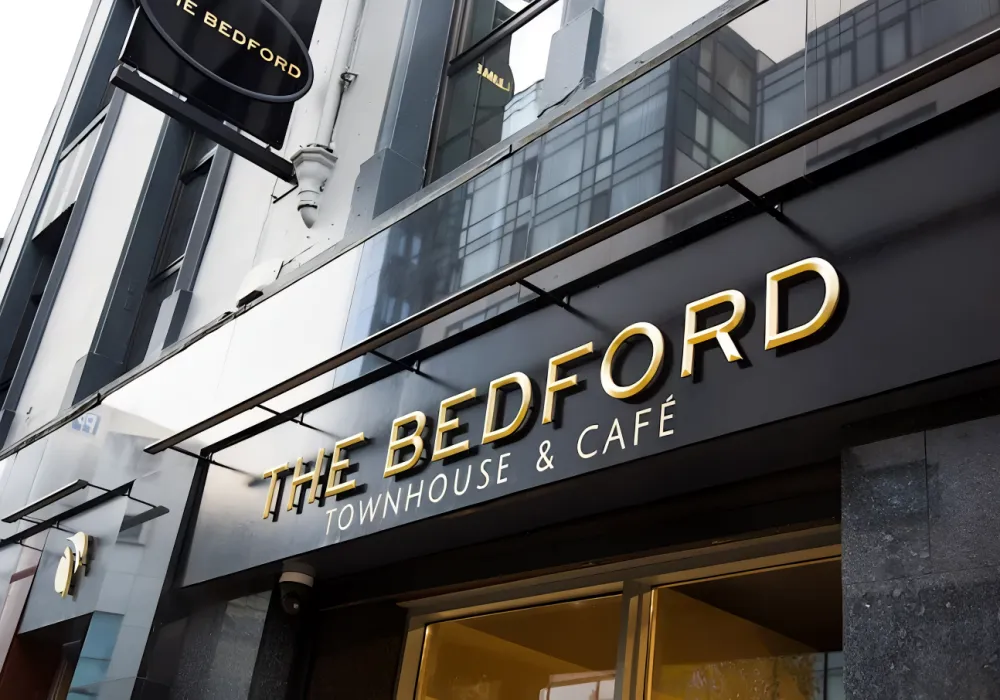 Gold letters spell out THE BEDFORD TOWNHOUSE & CAFÉ on a sleek black sign above a modern building entrance, with reflections of nearby buildings visible in the glass facade.